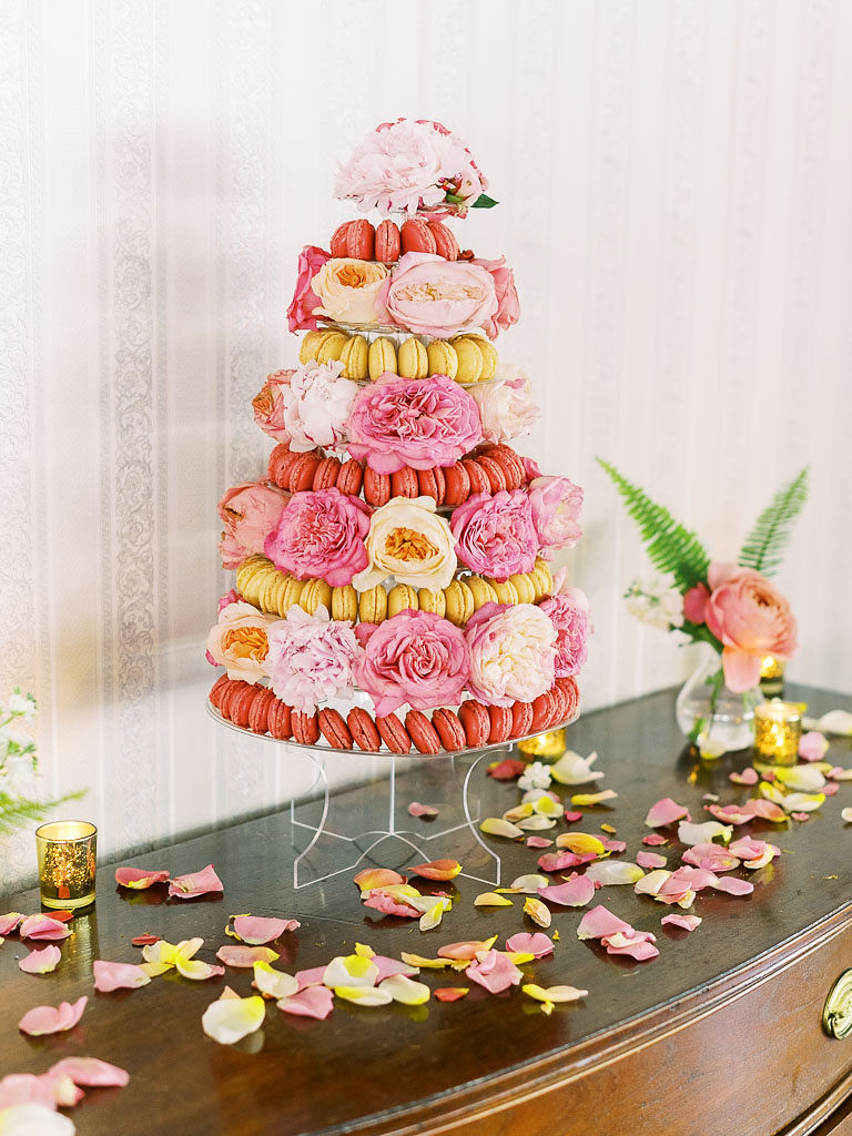 A tower of macarons on a dark wooden table with flower petals on it. There are large white and pink blooms in between the tiers of macarons.