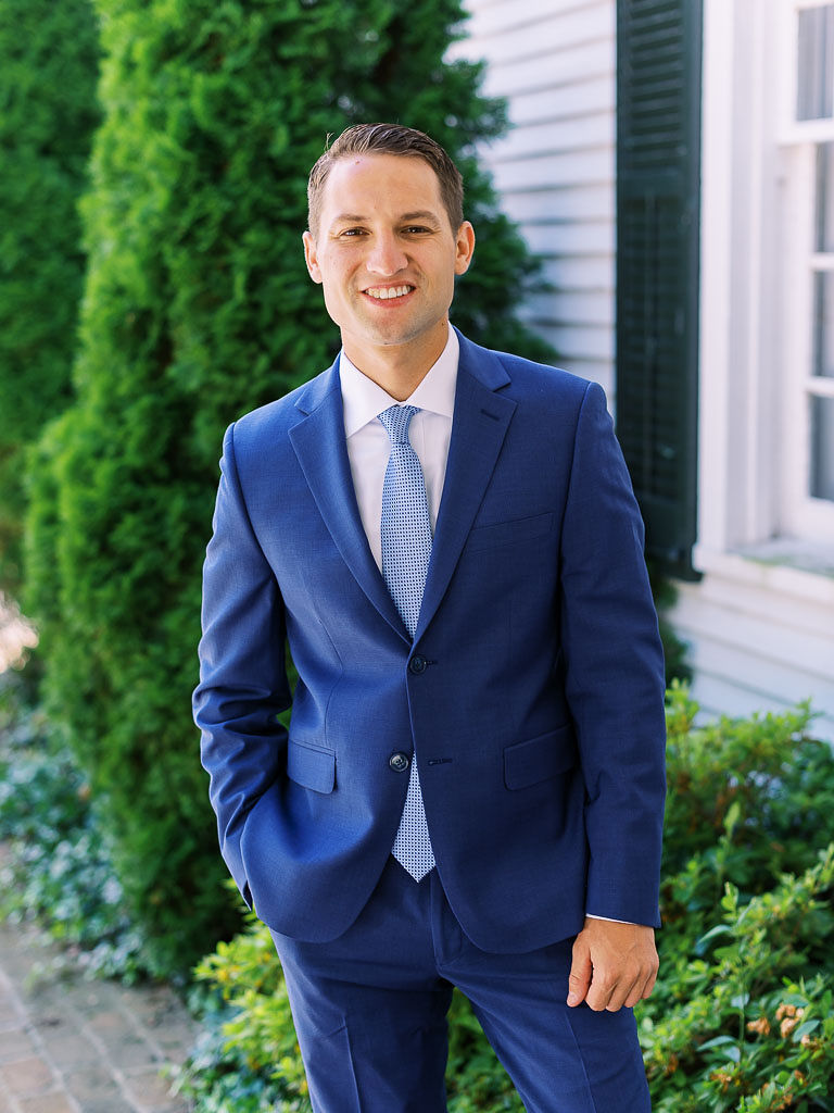 A groom on his wedding day standing next to a house, with his hand in his pocket. He is wearing a deep blue colored suit and light blue tie. He is looking at the camera and smiling.