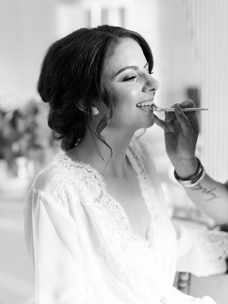 A black and white photo of a bride wearing a lace robe having her makeup done on her wedding day.