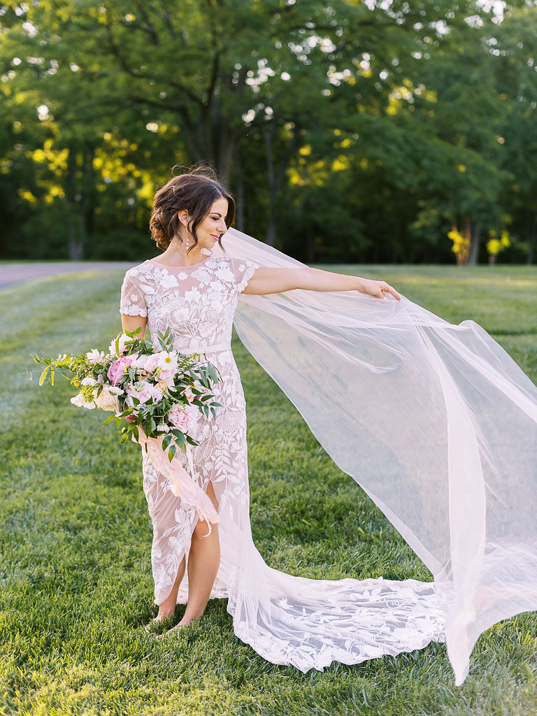 A bride standing on lush green grass with tall trees behind her. She is wearing her wedding gown is white lace flowers on it and nude-colored layer beneath that. Her long, sheer veil is blowing in the wind, and she is holding it. In her other hand is white and pink bouquet with cascading greenery. Taken by Maryland wedding photographer Kim Branagan at Antrim 1844.