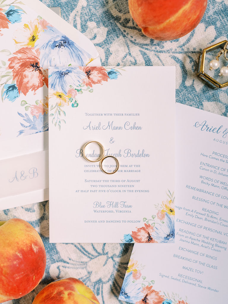Two gold wedding rings on top of a wedding invitation laying on a blue and white table cloth