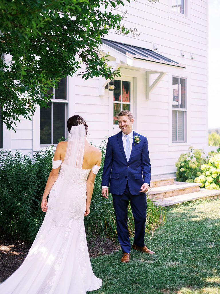 A bride and groom doing their first look on their wedding day. The bride is wearing a white wedding dress is a long veil, and the groom is wearing a blue suit,