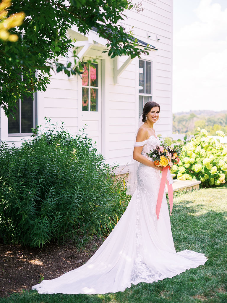 A bride stands outside at Blue Hill Farm in Virginia on her wedding day. She is wearing her wedding dress and holding a colorful bouquet.