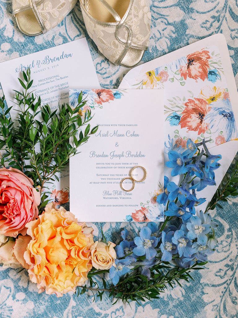 Two gold wedding rings on top of a wedding invitation laying on a blue and white table cloth , with peach, blue, and pink flowers surrounding the items. The bride's shoes are laying above the items.