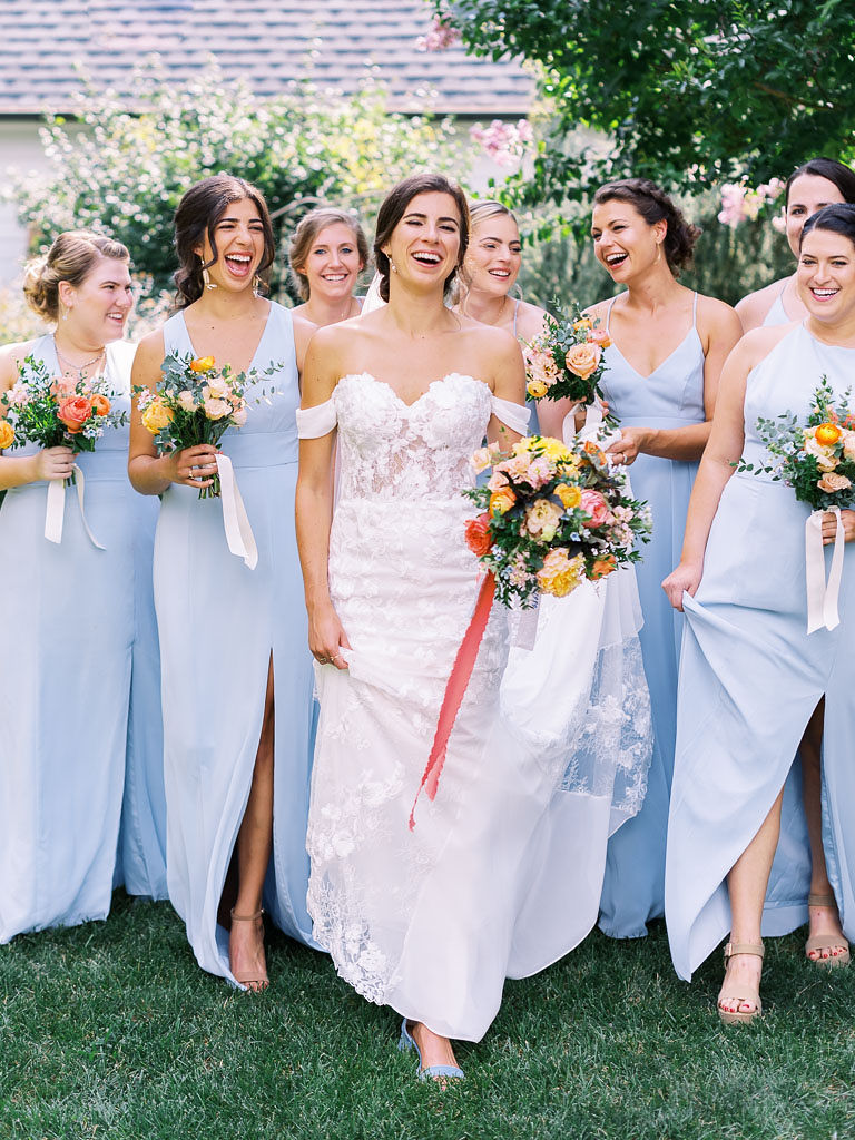 A bride and all her bridesmaids stand together smiling and laughing, holding their colorful floral bouquets
