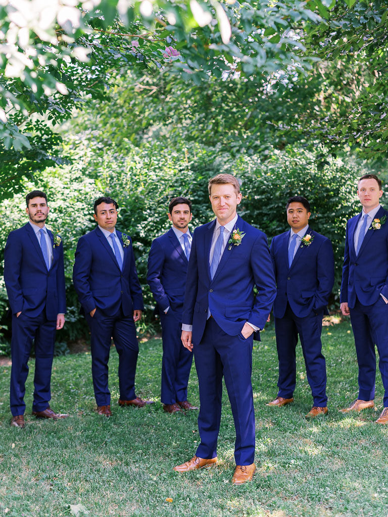 A man stands in front of his groomsmen outside; they are all smiling.