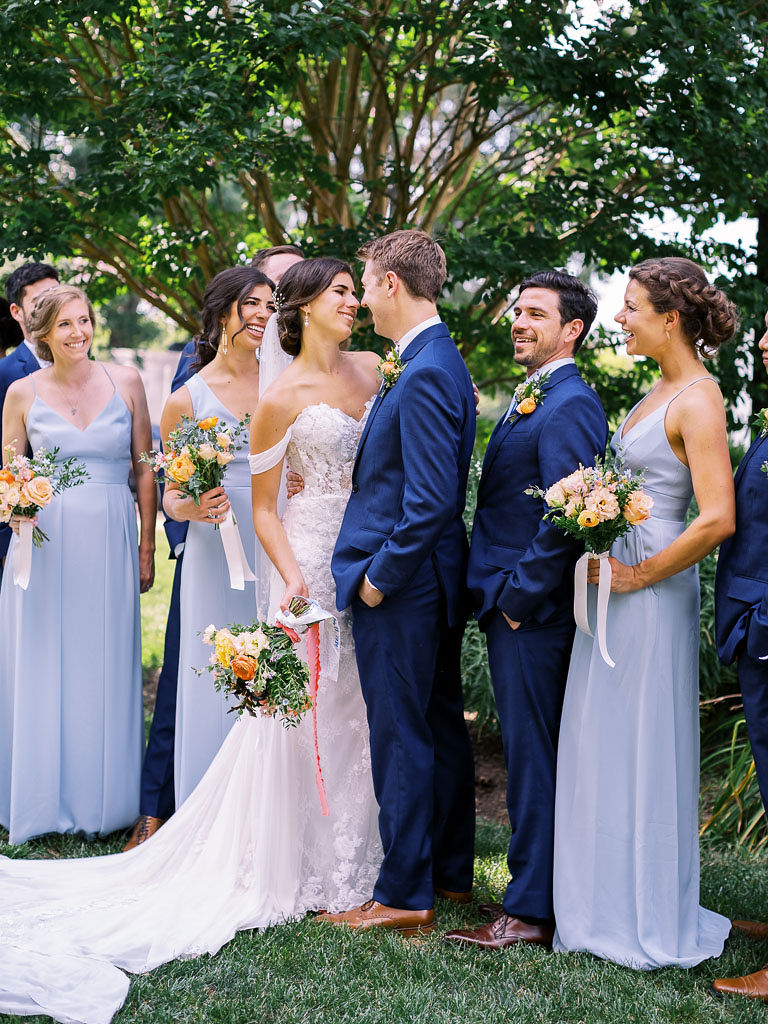 A bride and groom hold each other and gaze into each other's eyes with their wedding party surrounding them, smiling.