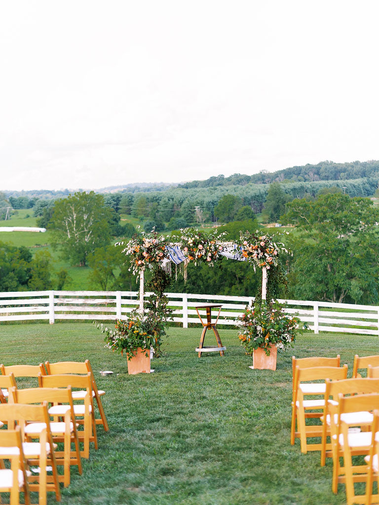 A lush floral archway set up in front of rolling hills for a wedding ceremony, with chairs in the foreground for the wedding guests. Photo by Virginia and Washington D.C. wedding photographer Kim Branagan.
