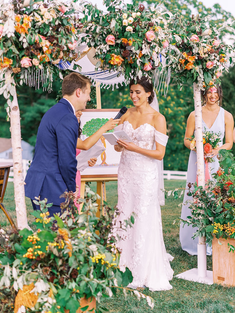 A bride reads her wedding vows to her groom during their wedding ceremony under a peach and blush floral archway. They are smiling at each other. Photo by Virginia and Washington D.C. wedding photographer Kim Branagan.