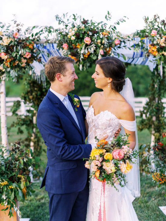 A couple in wedding attire holds each and smiles, standing in front of the floral arch they were just married under. Photo by Virginia wedding photographer Kim Branagan.