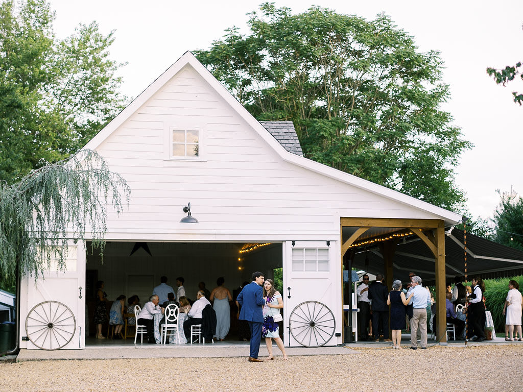 A white barn converted into a venue space at Blue Hill Farm in Waterford, VA. Wedding guests mingle in and around the barn. Photo by Virginia and Washington D.C. wedding photographer Kim Branagan.