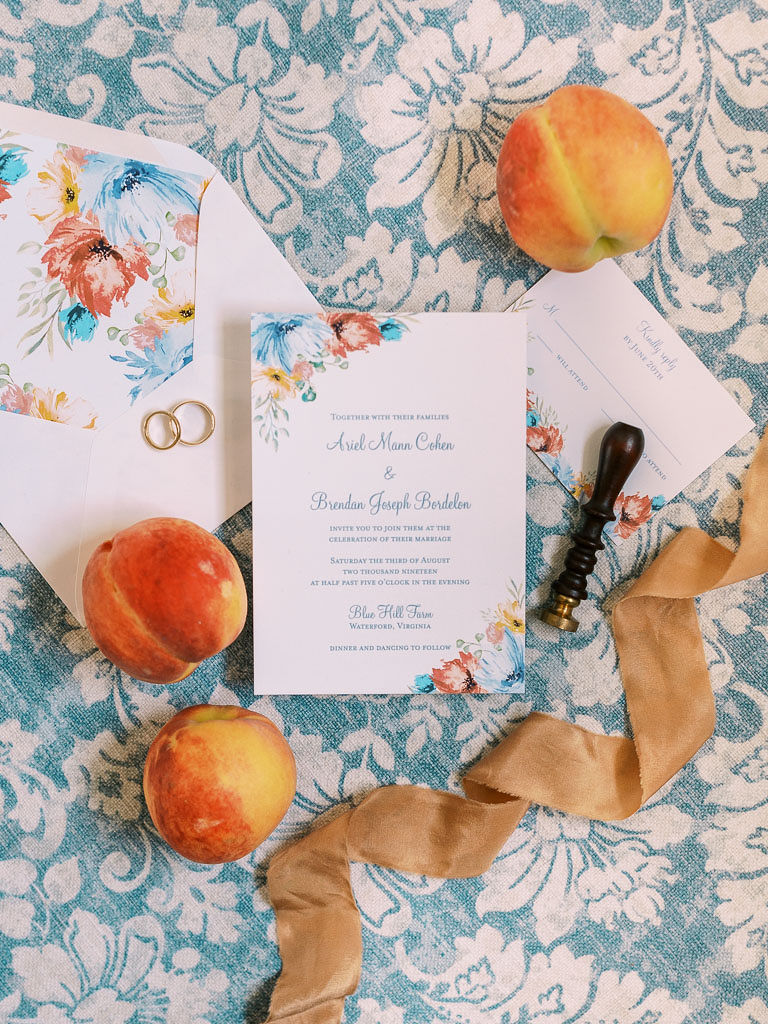 Save the date and wedding invitations on a blue and white floral tablecloth, which peaches and satin ribbon next to the papers. Photographer by Virginia wedding photographer Kim Branagan.