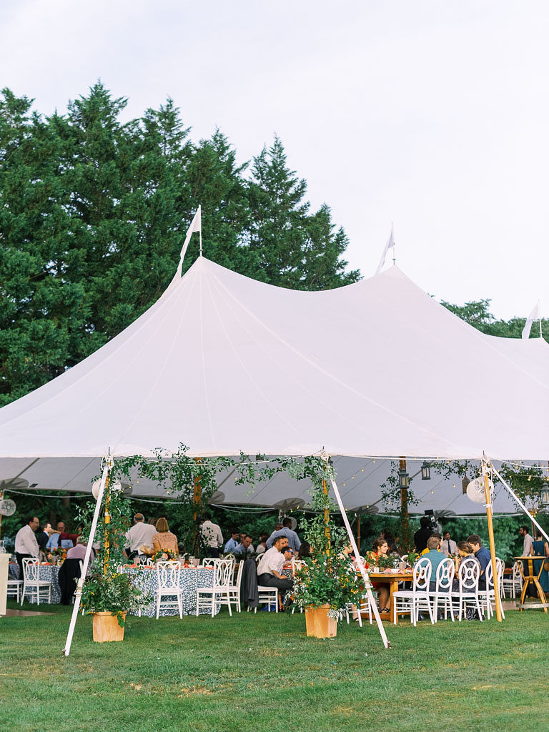 A large, white tent set up for an outdoor wedding reception, with wedding guests sitting down at tables under the tent.