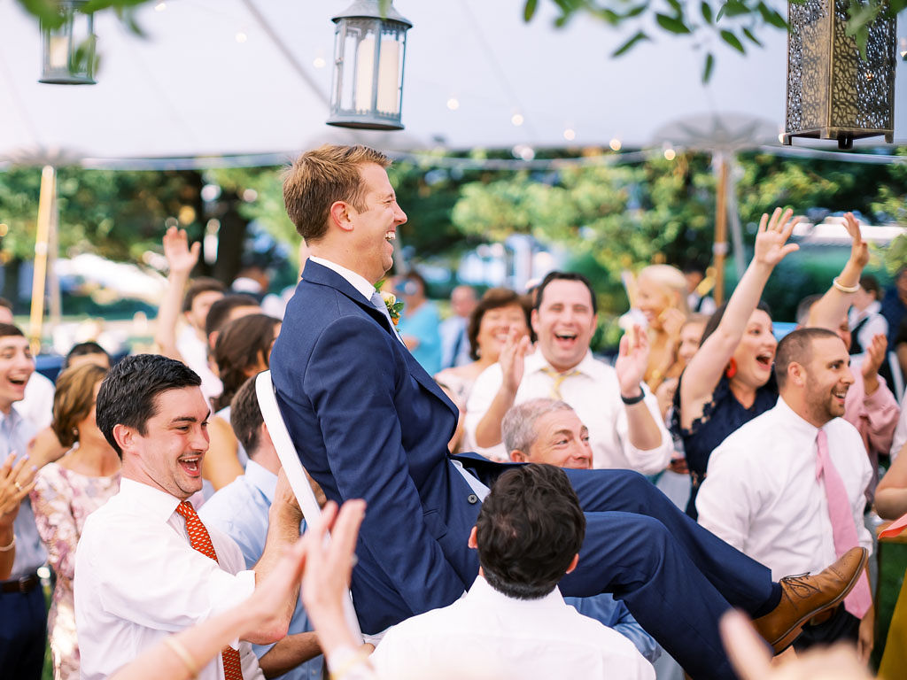A man sits on a chair as his groomsmen carry him on his wedding day. They are on the dance floor, and everyone is smiling and laughing.