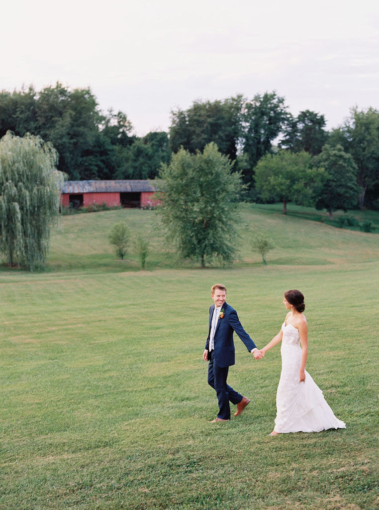 A bride and groom walk through a large green, hilly field. They are holding hands, looking at each other, and smiling. Photo by Virginia and Washington D.C. wedding photographer Kim Branagan.