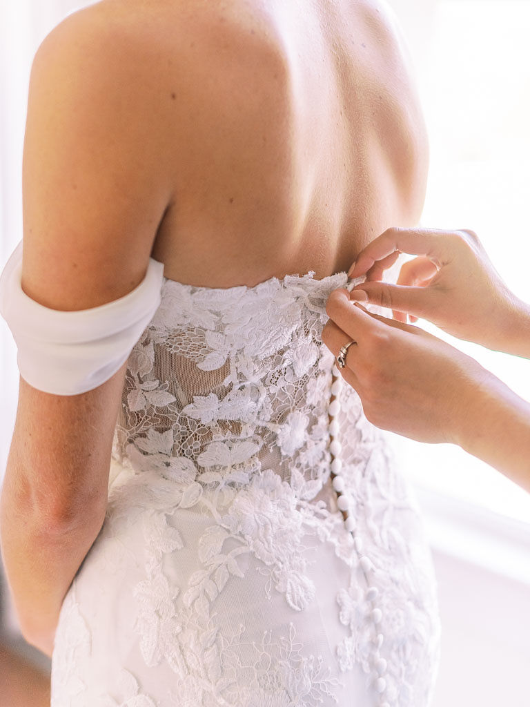 A bridesmaid buttoning up the back of the bride's white wedding gown
