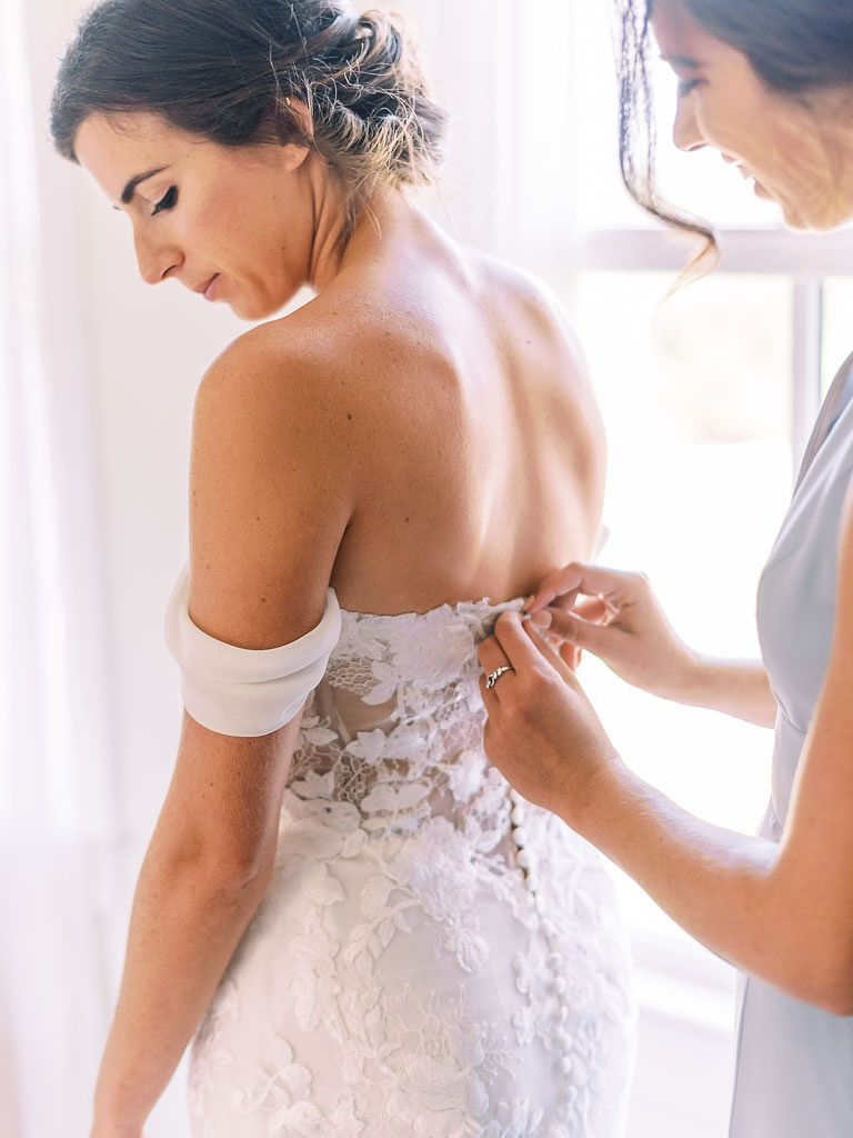 A smiling bridesmaid buttons up the bride's wedding dress as the bride looks down. Photographed by Washington D.C. wedding photographer Kim Branagan