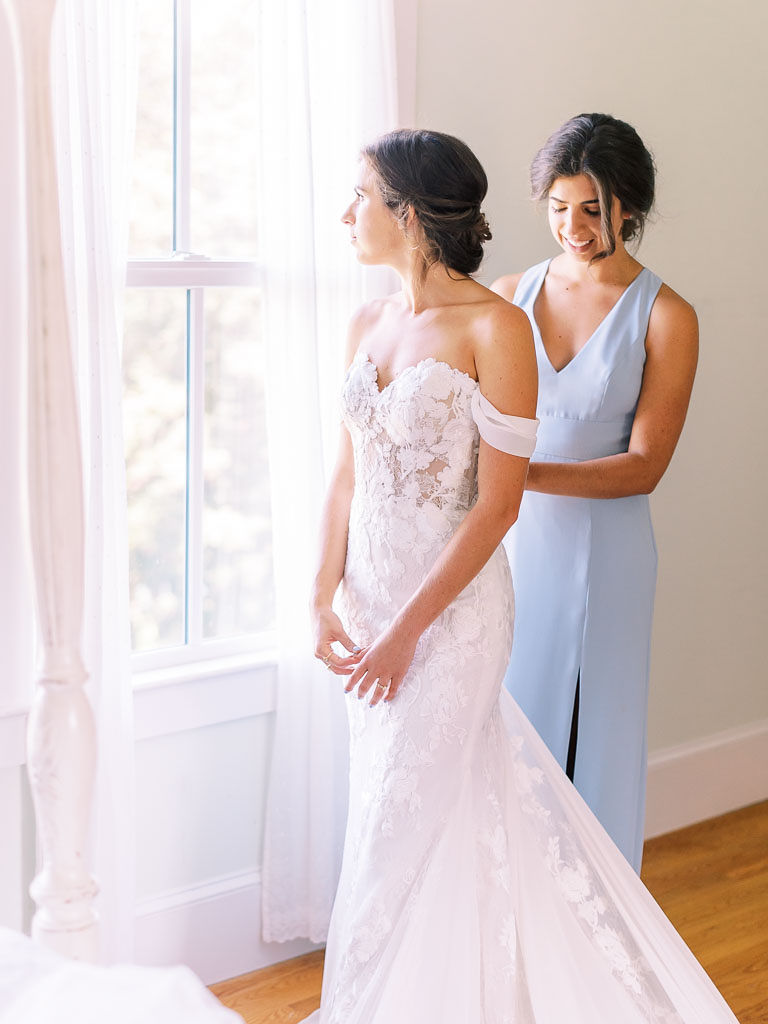 A bride looks out the window as one of her bridesmaids stands behind her and fixes her wedding dress