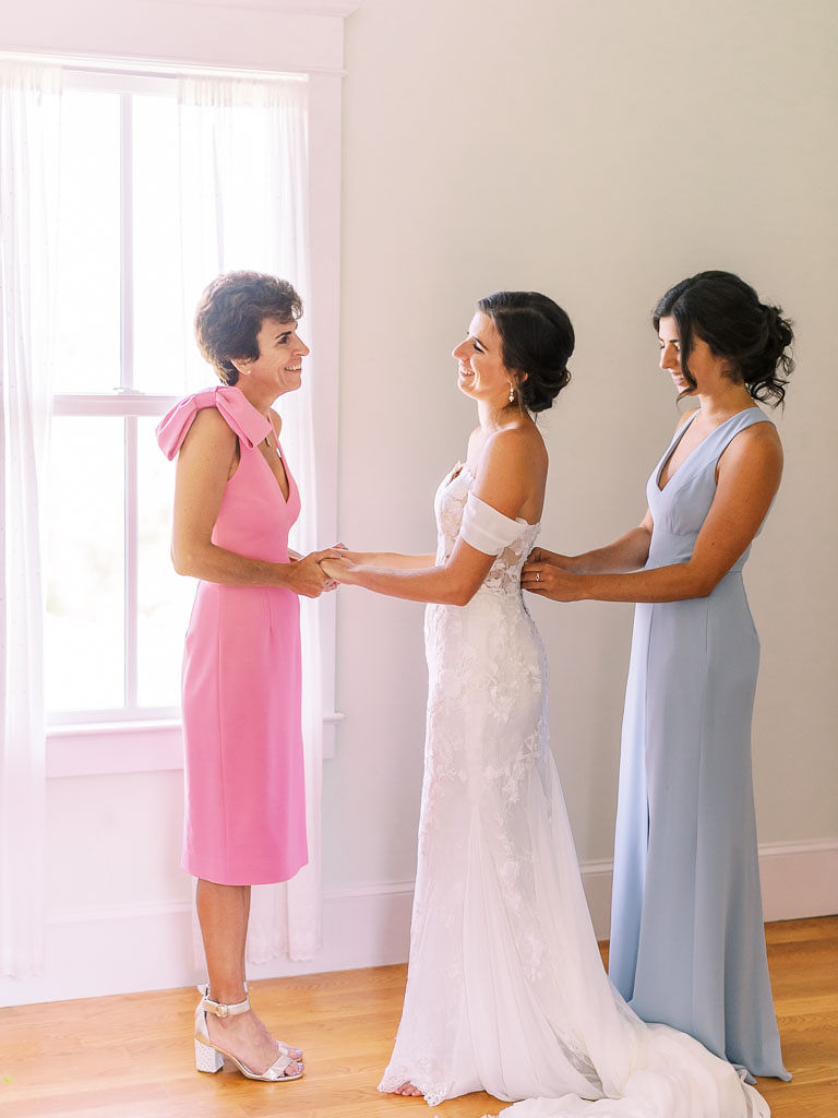 The bride's mother holds the bride's hands, and they smile at each other, as a bridesmaid buttons up the bride's dress