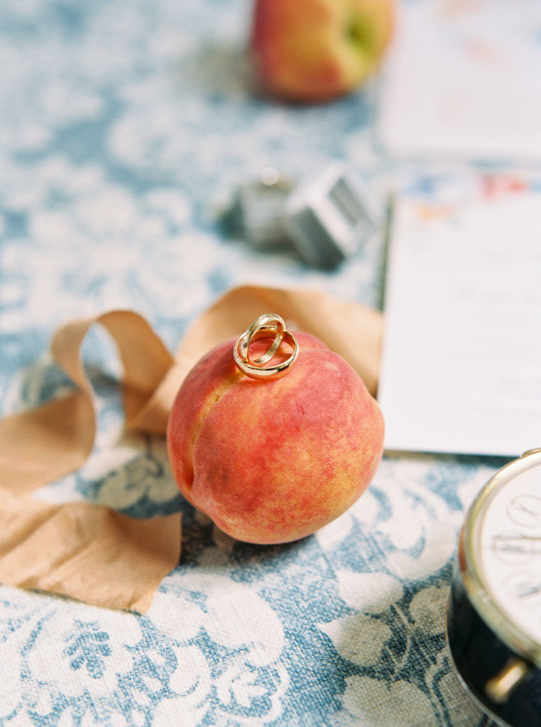 Two gold wedding rings on top of a peach on a blue and white table cloth.