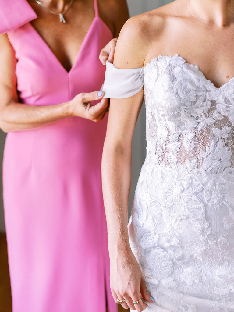 The bride's mother, wearing a pink dress, fixes the sleep of her daughter's wedding dress as her daughter stands in front of her