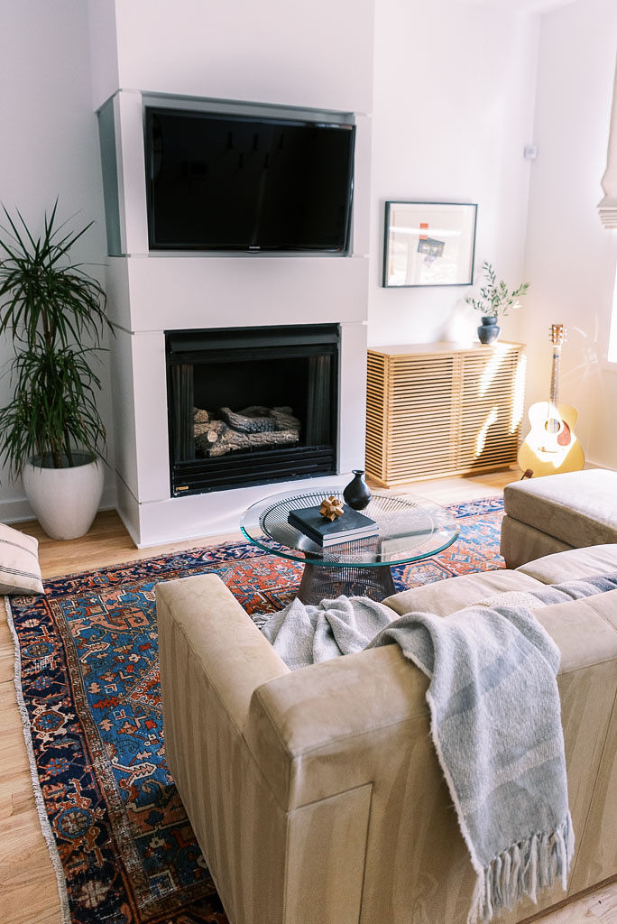 The living room of a minimalist, mid-century style Washington D.C. home. A large TV is mounted on the wall. There is a large plant on the ground to the left of the TV, and a colorful oriental rug covers the wood floor. A light gray, tasseled throw blanket from Parachute Home lays on the couch. Photographed by Washington DC photographer Kim Branagan.