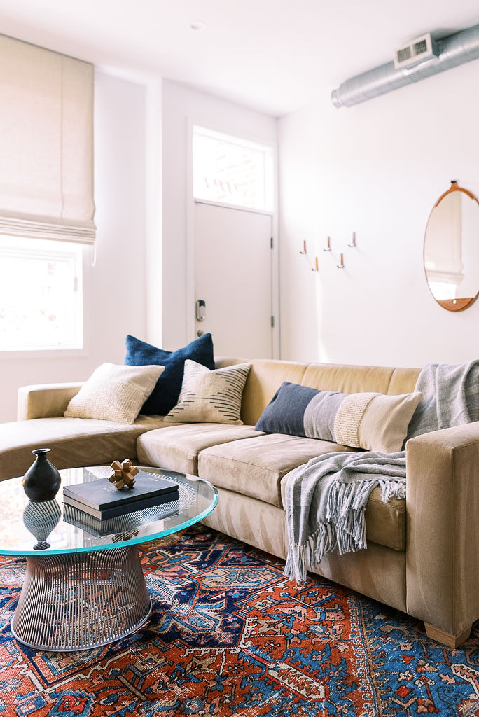 The living room of a minimalist, mid-century style Washington D.C. home. A colorful oriental rug covers the wood floor. A light gray, tasseled throw blanket from Parachute Home lays on the large, tan colored couch. Photographed by Washington DC photographer Kim Branagan.