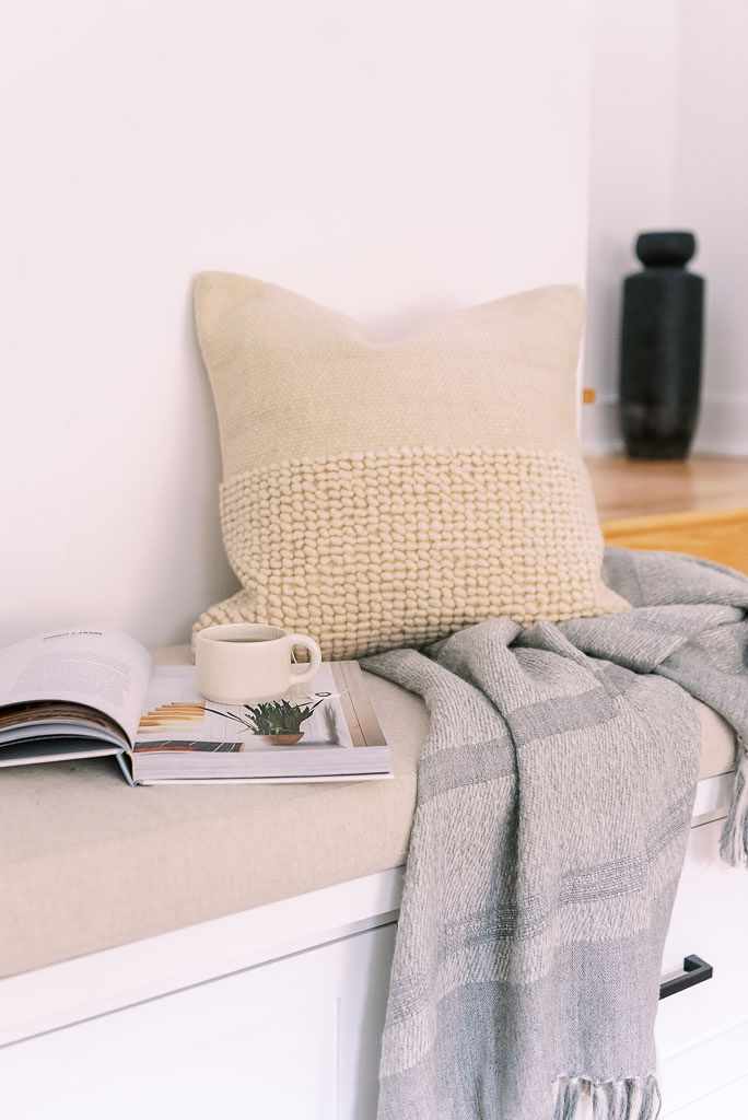 Close up shot of a cream throw pillow and gray throw blanket next to an open book and cup of tea on a bench in corner nook of a Washington D.C. home.