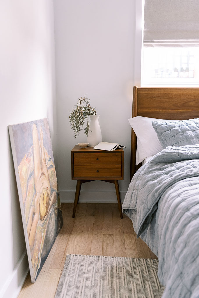 A mid century wooden nightstand next to a bed with light blue-gray bedding. A large painting leans against the wall opposite of the bed.