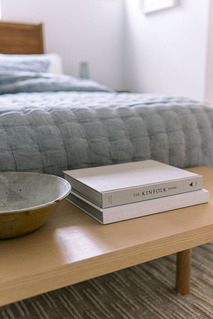 A small wooden bench at the foot of a bed with light gray bedding from Parachute Home. There are two books and a large bowl on the bench. Photographed by Washington DC commercial photographer Kim Branagan.