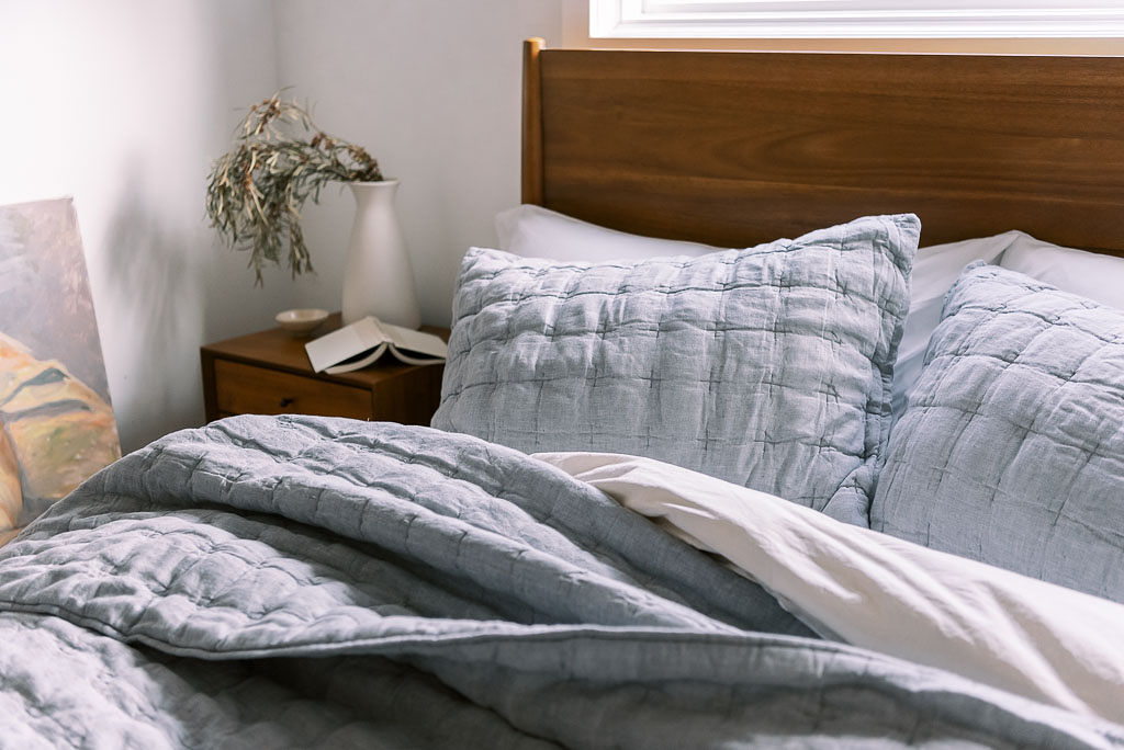 Close up shot of light blueish gray cotton bedding from Parachute Home on a bed against a wooden headboard in a Washington D.C. home. There is a small wooden nightstand with a green plant on it next to the bed.