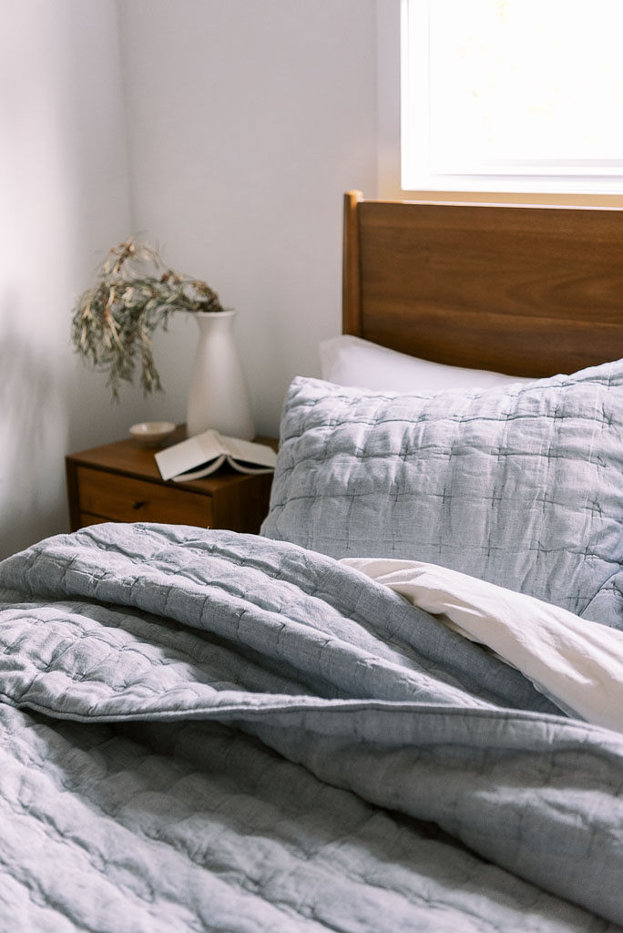 Close up shot of light blueish gray cotton bedding and white sheets from Parachute Home on a bed against a wooden headboard in a Washington D.C. home. There is a small wooden nightstand with a green plant on it next to the bed.