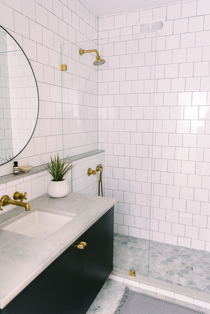 Photo of a bathroom of a bright white, modern style bathroom in a Washington D.C. home. The faucet and shower head are gold-colored. The walls and shower tiles are white. The floors are gray tiled, and the counter top is light marble with black cabinets below.