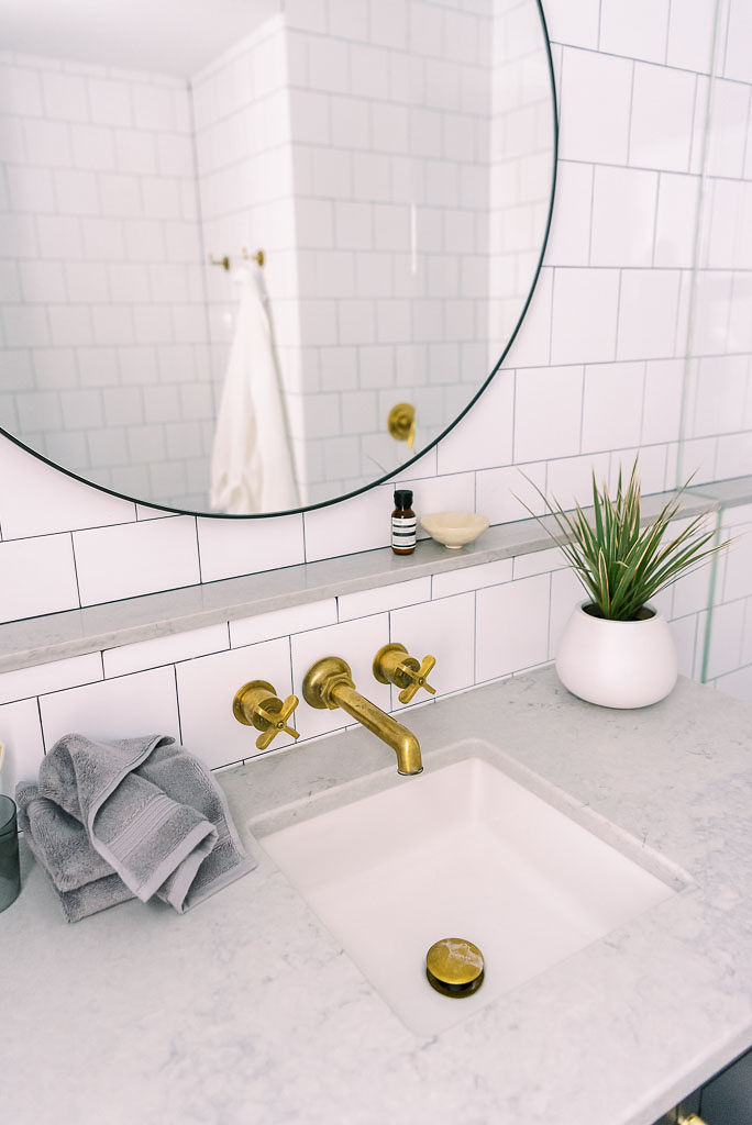 Close up shot of a bathroom sink, with gold knobs, a white marble counter top, and white tiled walls. There is a large round mirror hanging above the sink. A gray hand towel lays on the counter. Photographed by greater DMV area photographer Kim Branagan for Parachute Home.