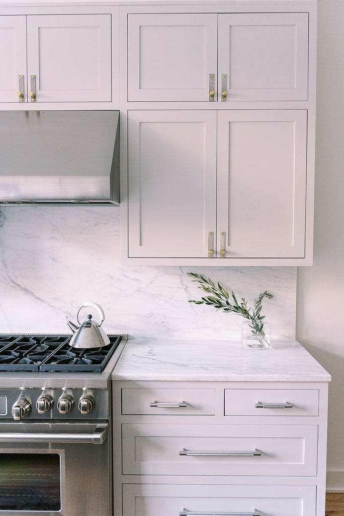 Close up shot of a stainless steel stove top with a tea kettle on it, against a white marble wall with white cabinets. A small olive branch in a vase is on the counter. Photographed by Washington D.C. photographer Kim Branagan for Parachute Home.