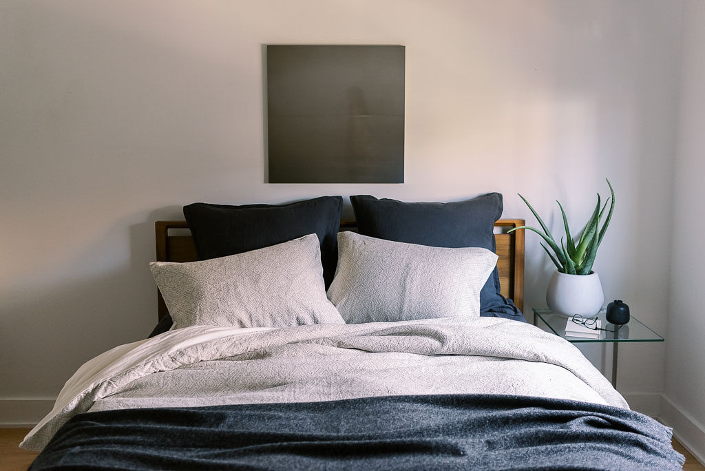 A navy bedspread and two navy pillows on a bed with light gray sheets and two light gray pillows against a wooden headboard. A charcoal painting hangs on the wall behind the bed. All bedding from Parachute Home, photographed by Kim Branagan.