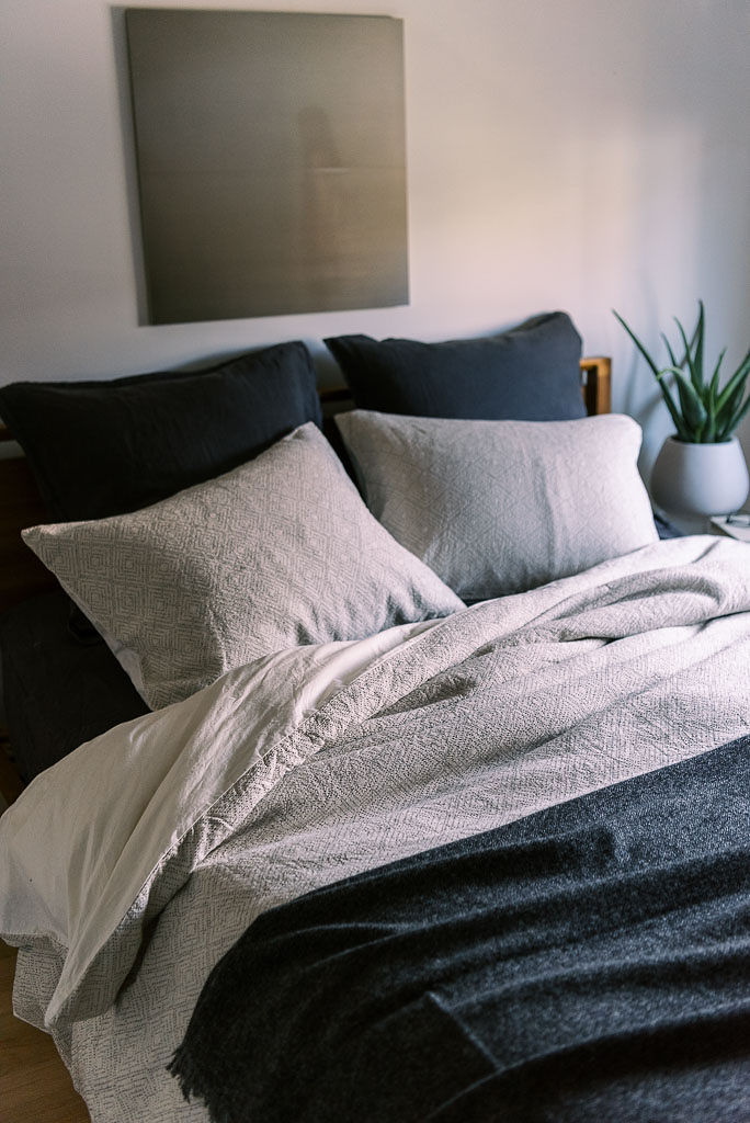 A texture navy bedspread and two navy pillows on a bed with textured light gray sheets and two light gray pillows against a wooden headboard. A charcoal painting hangs on the wall behind the bed. All bedding from Parachute Home, photographed by Kim Branagan.