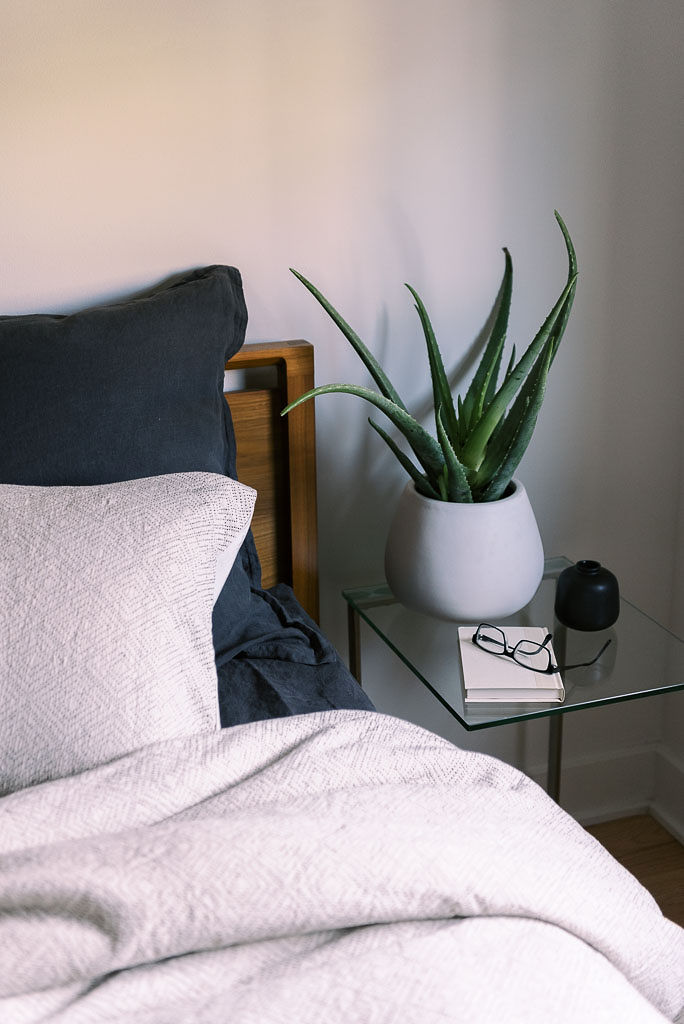 Close up shot of a glass nightstand with an aloe plant on it next to a bed with white-gray sheets and a navy pillow.