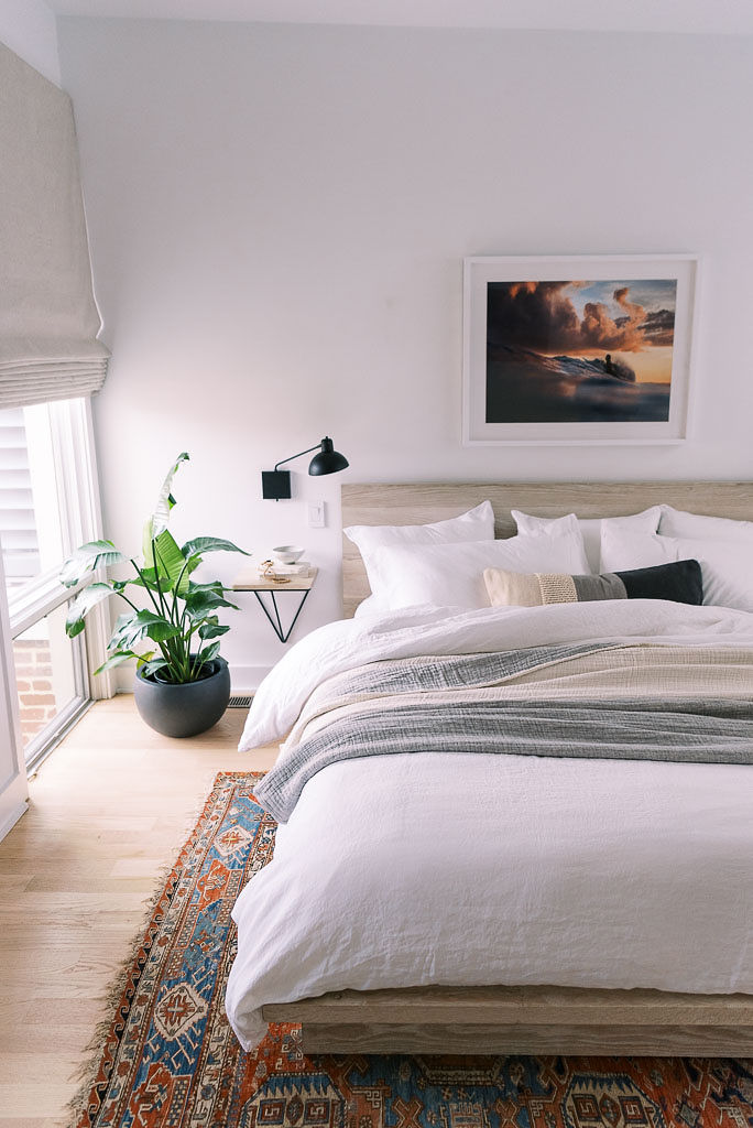 Photo of a mid century, boho style bedroom in a Washington D.C. home. The bedding is white linen with a gray throw blanket and gray and cream throw pillow on it, all from Parachute Home,against a light wood headboard. A colorful oriental rug covers the wood floor under the bed. There is a floor to ceiling window in the room, giving lots of natural light to the white colored walls. There is a large green plant on the ground by the window. Photographed by Washington DC commercial and interior photographer Kim Branagan.