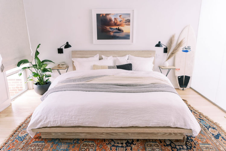Photo of a mid century, boho style bedroom in a Washington D.C. home. The bedding is white linen with a gray throw blanket and gray and cream throw pillow on it, all from Parachute Home, against a light wood headboard. A colorful oriental rug covers the wood floor under the bed. There is a floor to ceiling window in the room, giving lots of natural light to the white colored walls. There is a large green plant on the ground by the window. A small nightstand with a lamp on it is next to the bed. There is a white surfboard on the other side of the nightstand. Photographed by Washington DC commercial and interior photographer Kim Branagan.