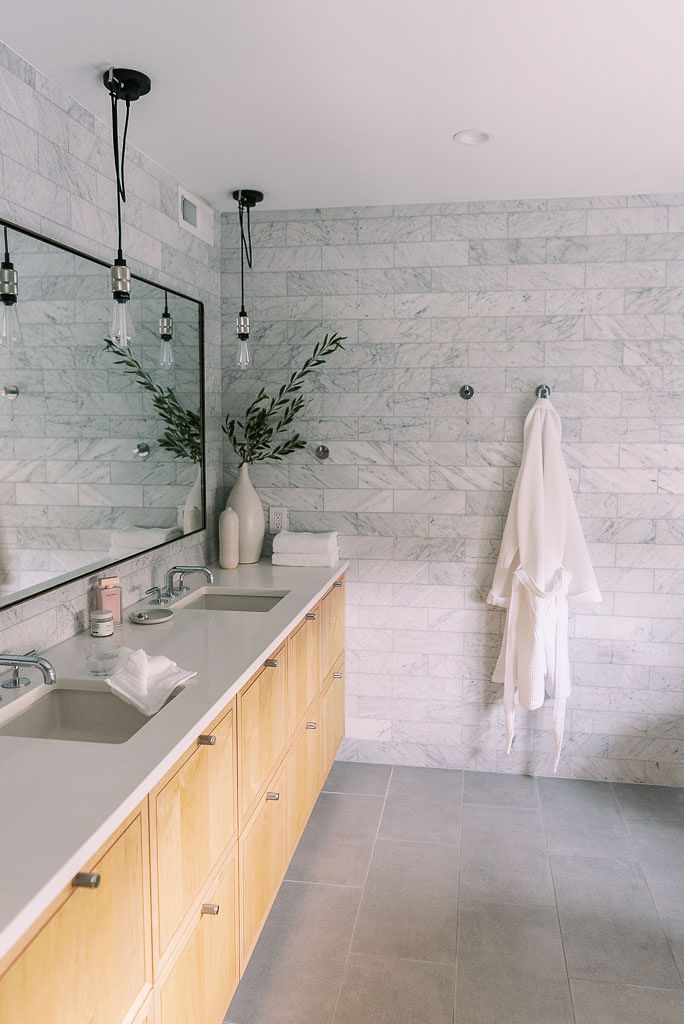 Photo of a minimalist style bathroom in a Washington DC home. A white robe hangs on one of the gray tiled walls, and a large rectangular mirror is on another wall. The floors are a darker gray colored tile. The counters are white and light colored wooden cabinets are below.