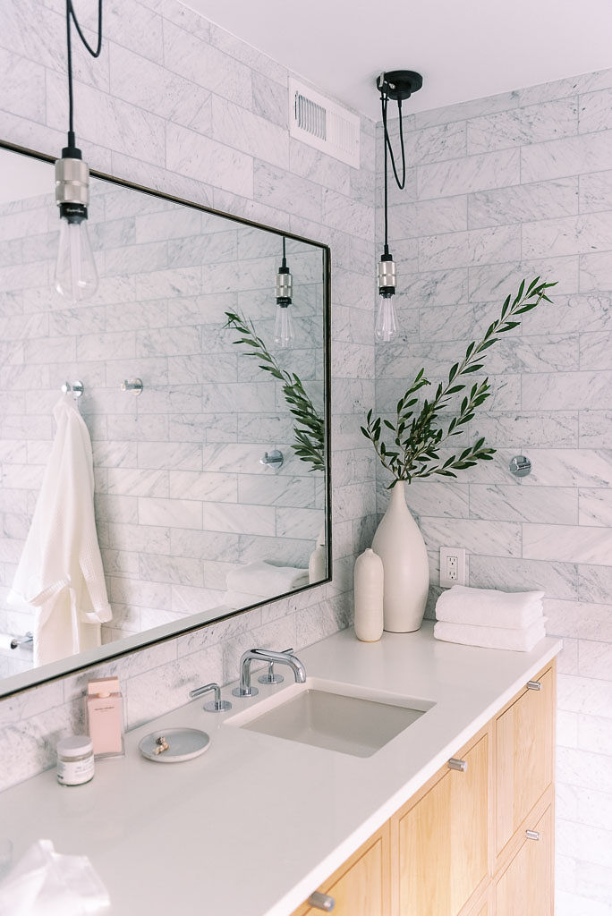 Photo of a minimalist style bathroom in a Washington DC home. a large rectangular mirror is on the light gray tiled wall. The counter is white and light colored wooden cabinets are below. Various toiletries are on the counter, as well as a tall green plant and two white towels from Parachute Home. Photographed by Washington DC interior design photographer Kim Branagan