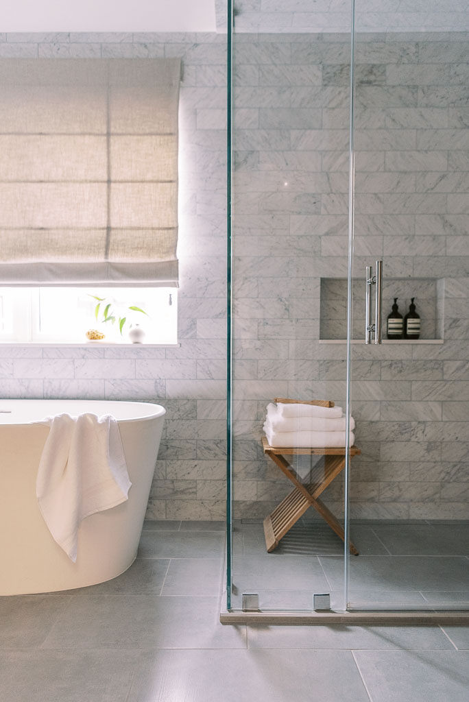 Photo of a minimalist, modern inspired bathroom in a Washington DC home. The shower is enclosed by clear glass. There is a small wooden stand with two white towels in the shower. The walls are a light gray marble, and the floor is a darker gray tile. There is a large white bathtub next to the shower area, with a white towel draping over the edge. There is a window with the shade mostly pulled down behind the tub. All towels are from Parachute Home. Photographed by Washington DC interior photographer Kim Branagan.