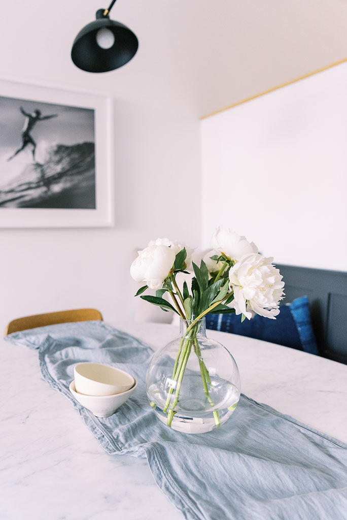 Close up shot of a white wooden table with a light blue table runner from Parachute Home. There is a vase of white flowers and two small bowls on the table runner.