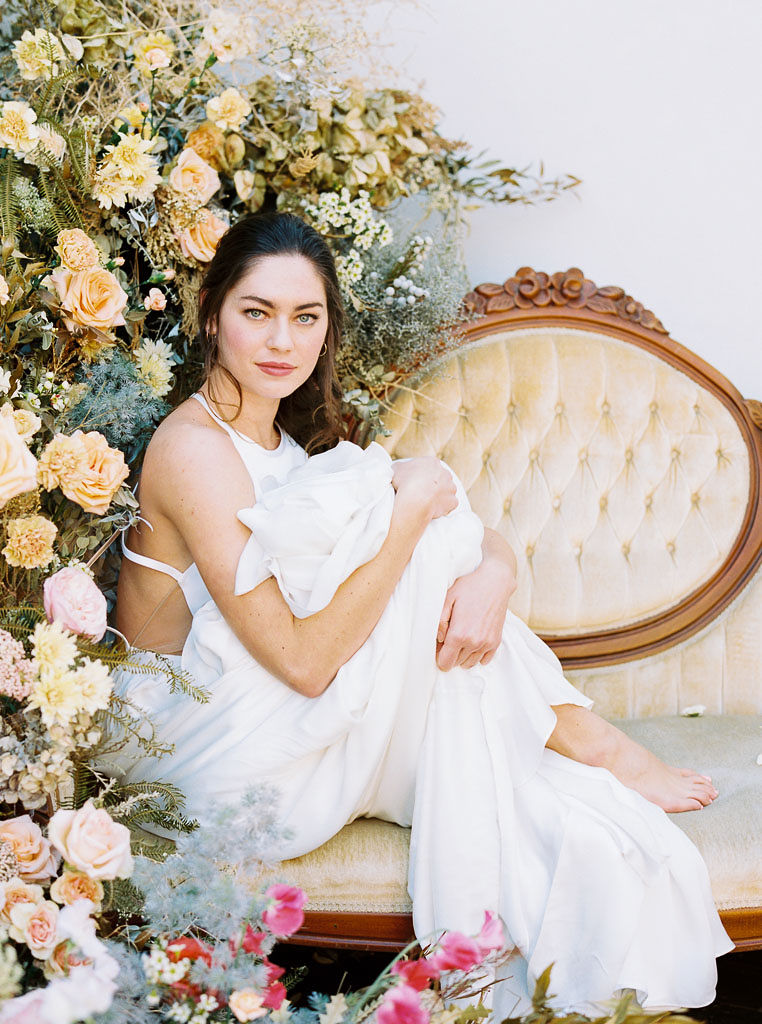 A model wearing a flowing white wedding dress sitting on a vintage velvet couch, next to a floral installation of yellow, blue, cream, and pink wildflowers. Photographed by San Diego editorial photographer Kim Branagan.