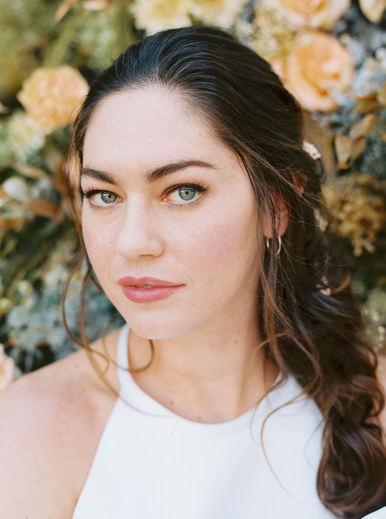 Close up shot of a woman with long brunette hair in a loose braid and her makeup professionally done staring straight at the camera. There are yellow and blue flowers behind her.