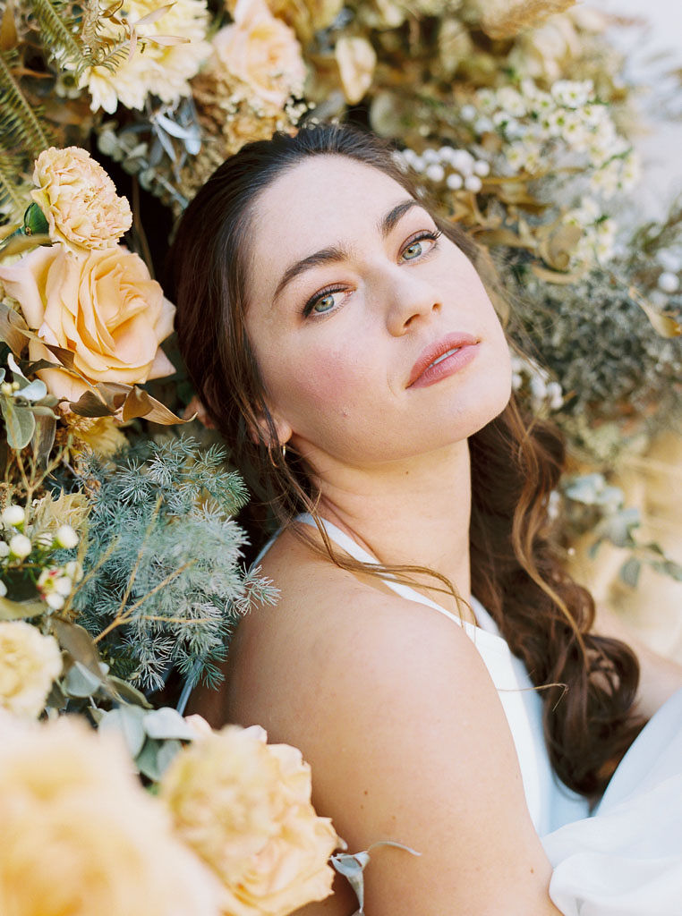 Close up shot of a woman with long brunette hair in a loose braid and her makeup professionally done staring straight at the camera. She is leaning against a large floral arrangement of yellow, white, and blue flowers designed by Nectar + Bloom floral studio. Photographed by San Diego commercial photographer Kim Branagan.