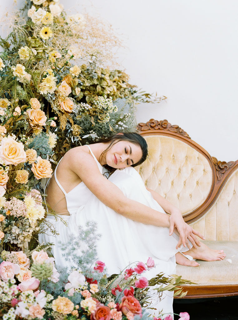 A large floral arrangement made of lush wildflower blooms and vibrant spring colors, surrounding an antique, yellow velvet sofa. A model wearing a flow-y, white wedding dress sits on the sofa, holding her knees to her chest and smiling. Photographed by Kim Branagan at Nectar + Bloom in San Diego, CA.