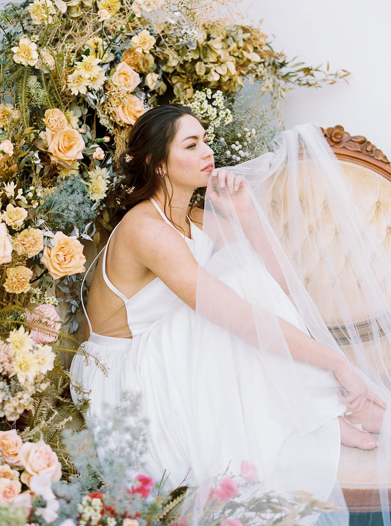 A large floral arrangement made of lush wildflower blooms and vibrant spring colors, surrounding an antique, yellow velvet sofa. A model wearing a flow-y, white wedding dress sits on the sofa, with tulle from the dress blowing in the wind. Photographed by Kim Branagan at Nectar + Bloom in San Diego, CA.