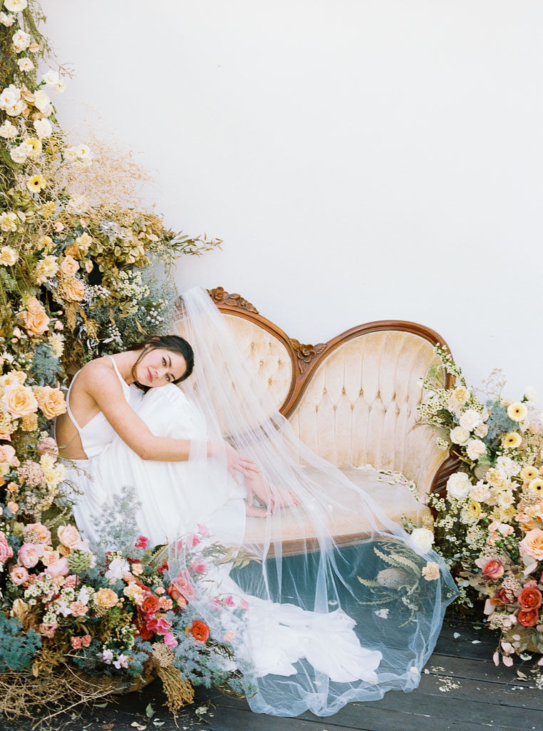 A large floral arrangement made of lush wildflower blooms and vibrant spring colors, surrounding an antique, yellow velvet sofa. A model wearing a flow-y, white wedding dress sits on the sofa. She is holding her knees to her chest, with her cheek resting on her knees and smiling. Photographed by Kim Branagan at Nectar + Bloom in San Diego, CA.